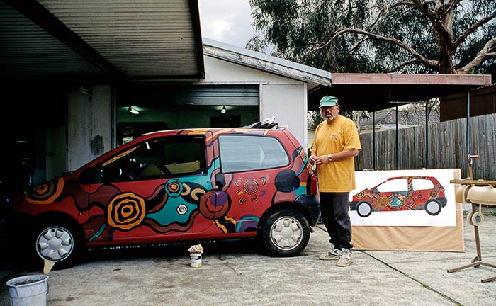 A painter works on the car in a backyard with the drawing next to him.