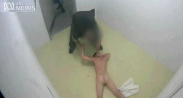 A teenager lies naked on the floor of his cell, his neck held down by a prison guard.
